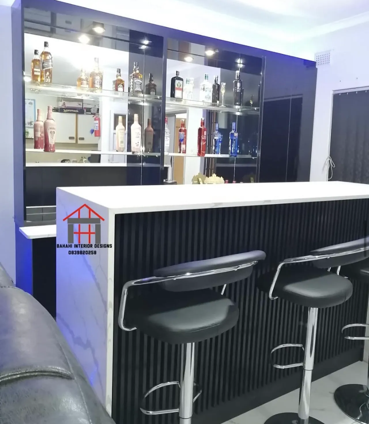 A home bar with a slatted black front, a white stone top, chrome bar stools and a mirrored, lit back wall with glass shelves.