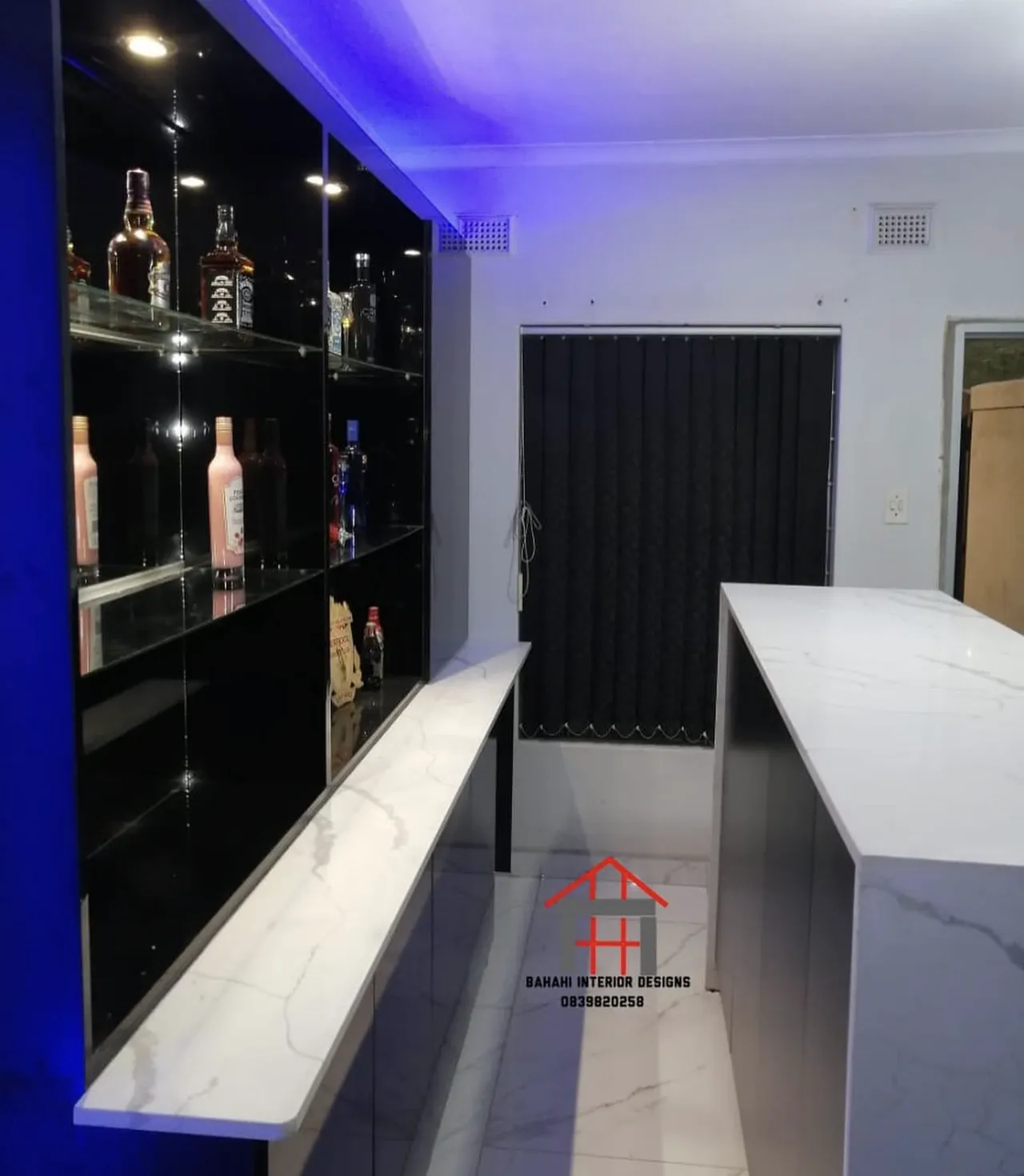 Behind the home bar: black gloss cabinets, glass shelves of bottles and a white stone back counter under blue light.