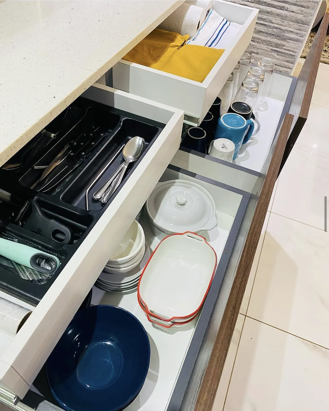 Deep pull-out drawers open under a white quartz top, holding cutlery, crockery and glasses.