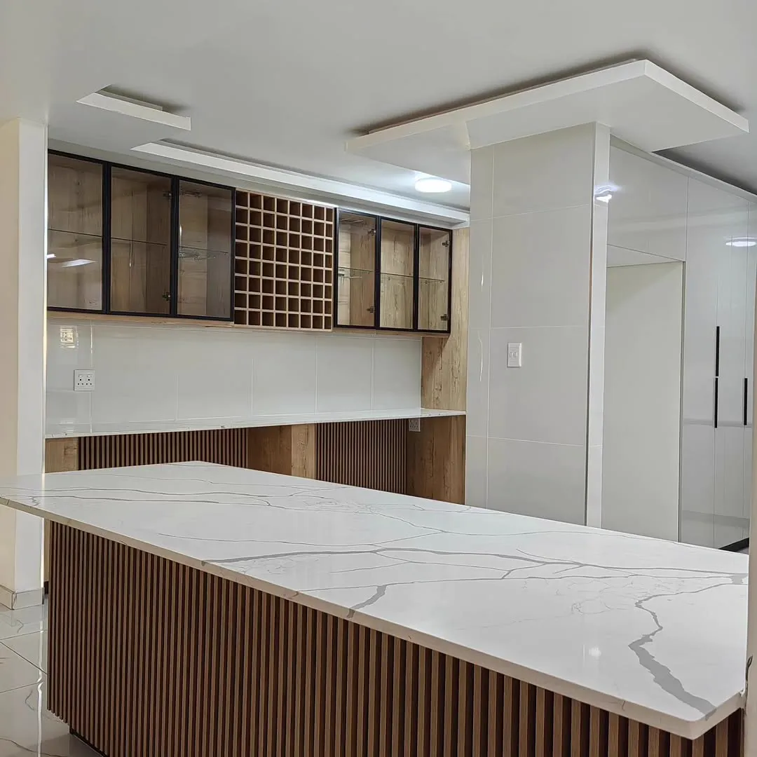 A long island with a veined white top and a timber-slat front, under black-framed glass cabinets and a wine rack.
