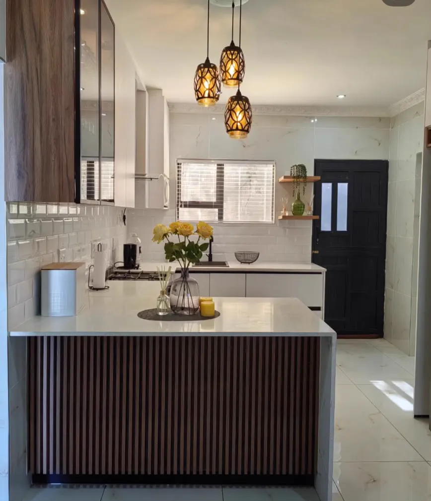 A white melamine kitchen with a timber-slat peninsula, brass pendant lights and subway tiles.