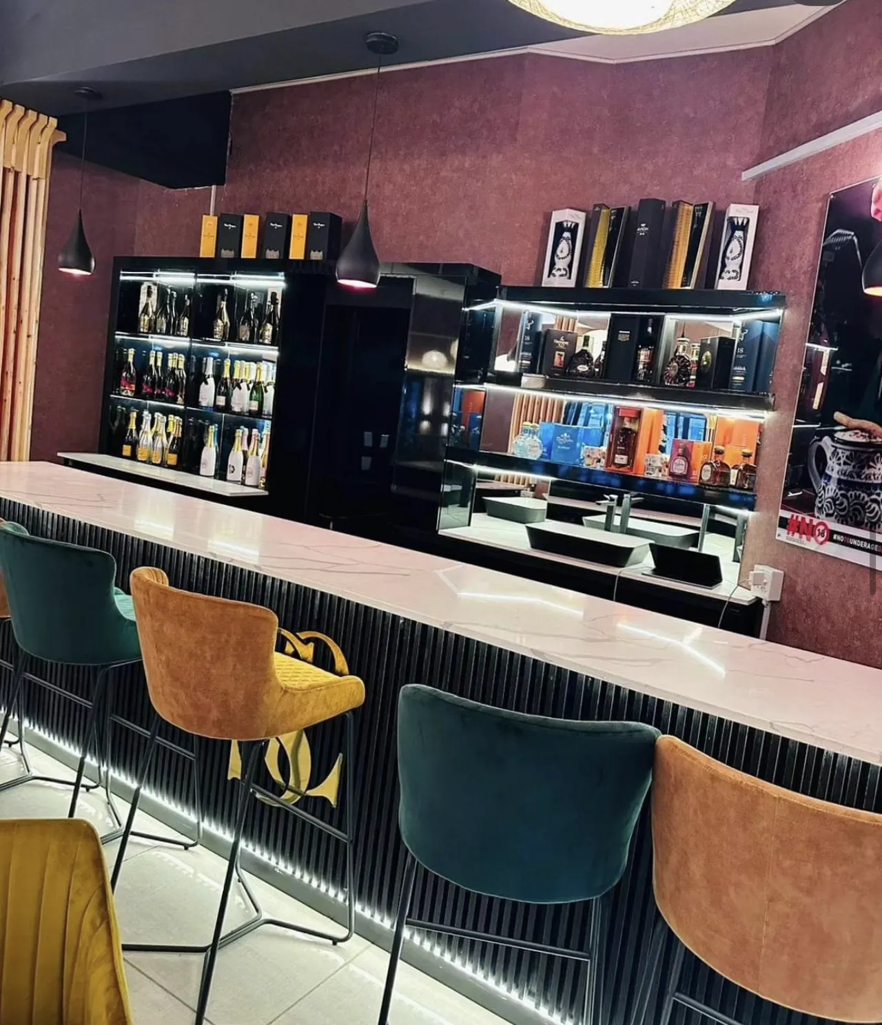 A restaurant bar with a dark slatted front lit from below, a veined stone counter, velvet stools and lit bottle shelving.