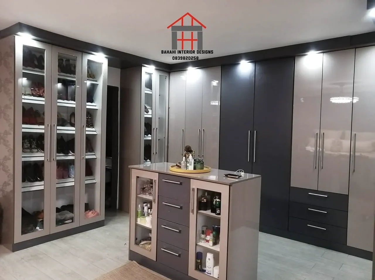 A walk-in closet in metallic cappuccino and storm grey gloss, with glass-fronted shoe towers and a centre island of drawers.